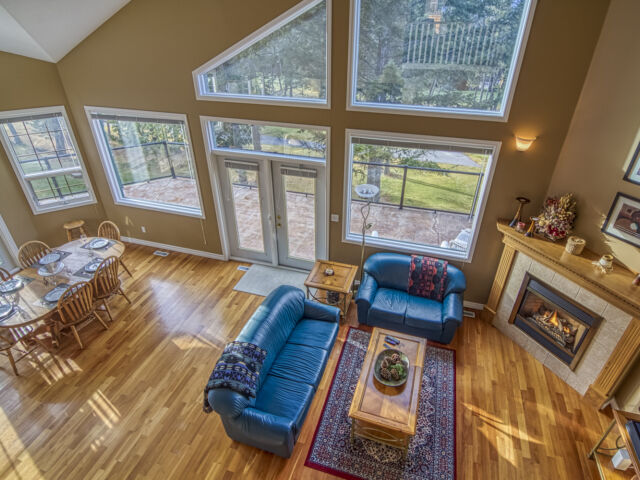 Dining room and living room as viewed from the loft above. Light wood dining table and chairs, blue couch and chair, rug, coffee table, and fireplace.